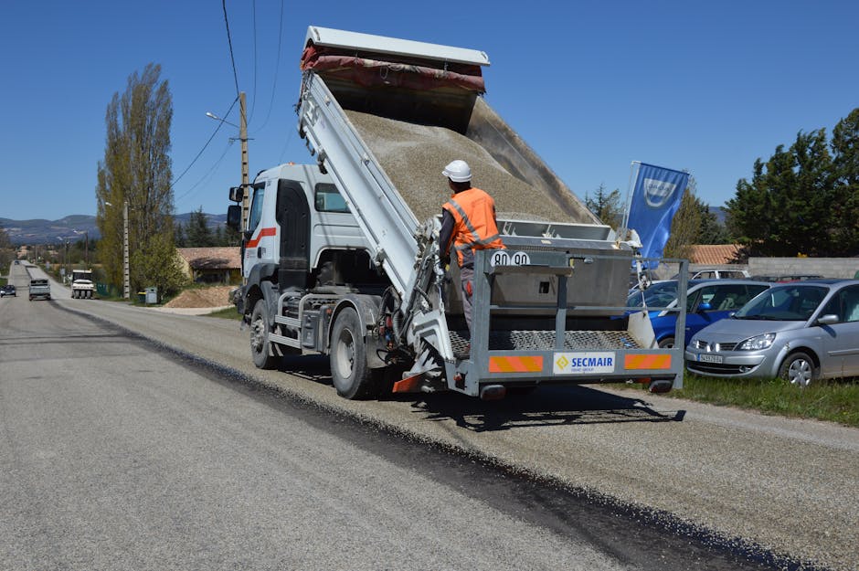 A dump truck unloading asphalt during road work with a worker in a reflective jacket.